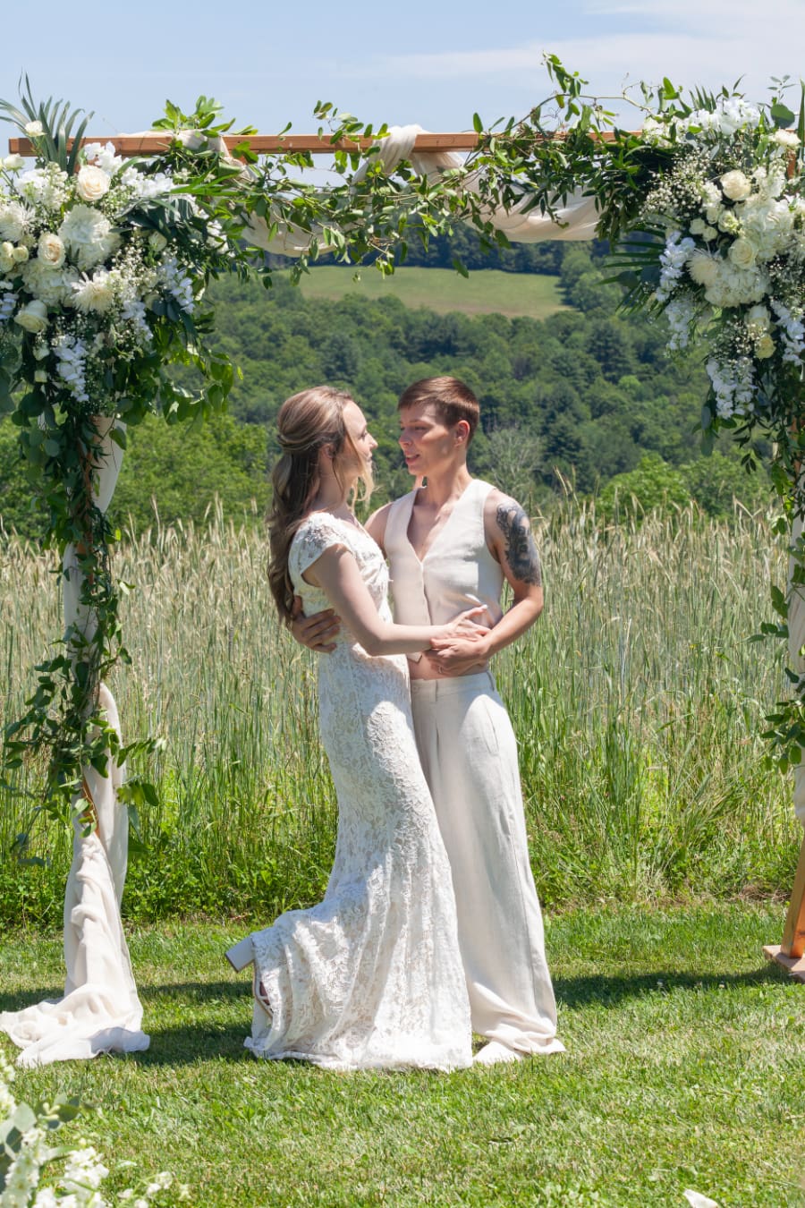 Couple under floral arch in a sunny field, sharing a moment during their rustic barn wedding.