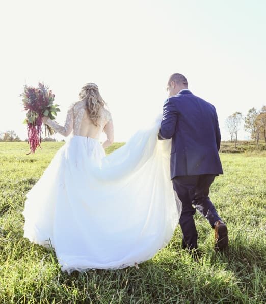 Bride and groom walking through a sunlit field, holding hands during their romantic barn wedding.