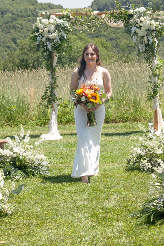A bride holding a vibrant bouquet stands beneath a floral arch in the garden at The Barn on Hubbard during her wedding weekend in the Catskills.