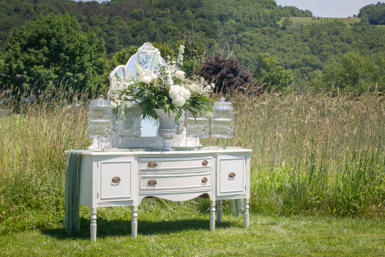A vintage white buffet table styled with florals and drink dispensers set in a meadow at The Barn on Hubbard, perfect for a wedding weekend in the Catskills.