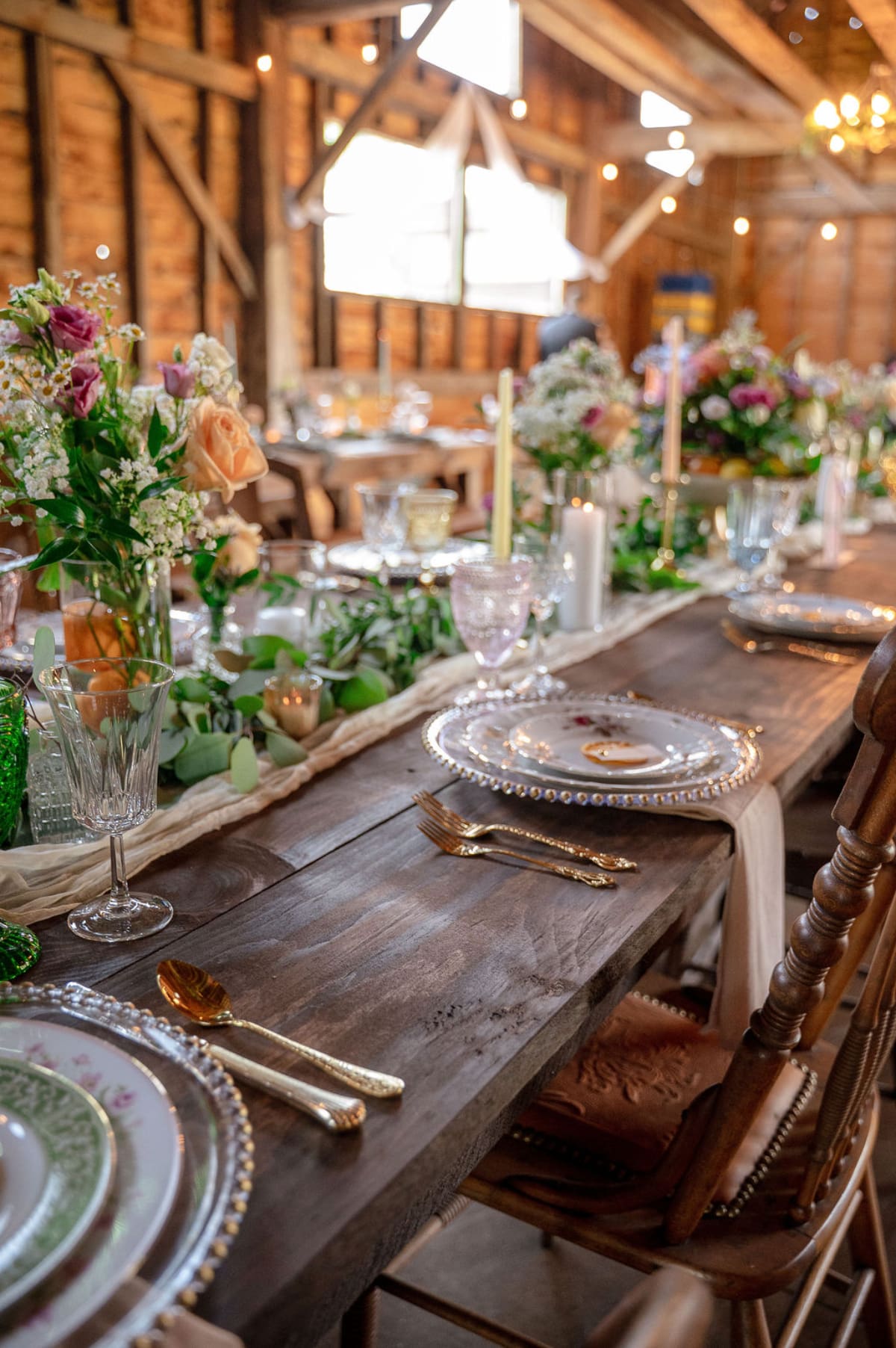 Long farmhouse table inside a restored barn with greenery, candles, and mismatched glassware, demonstrating elegant tablescapes often seen at affordable wedding venues.