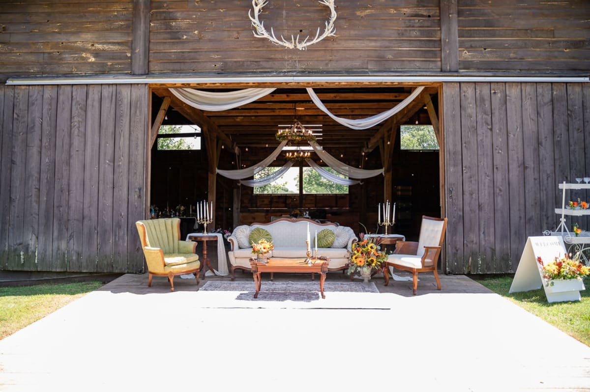 Open barn doors framing a styled seating vignette with draped fabric and soft lighting, showcasing how affordable wedding venues create welcoming ceremony or cocktail spaces.