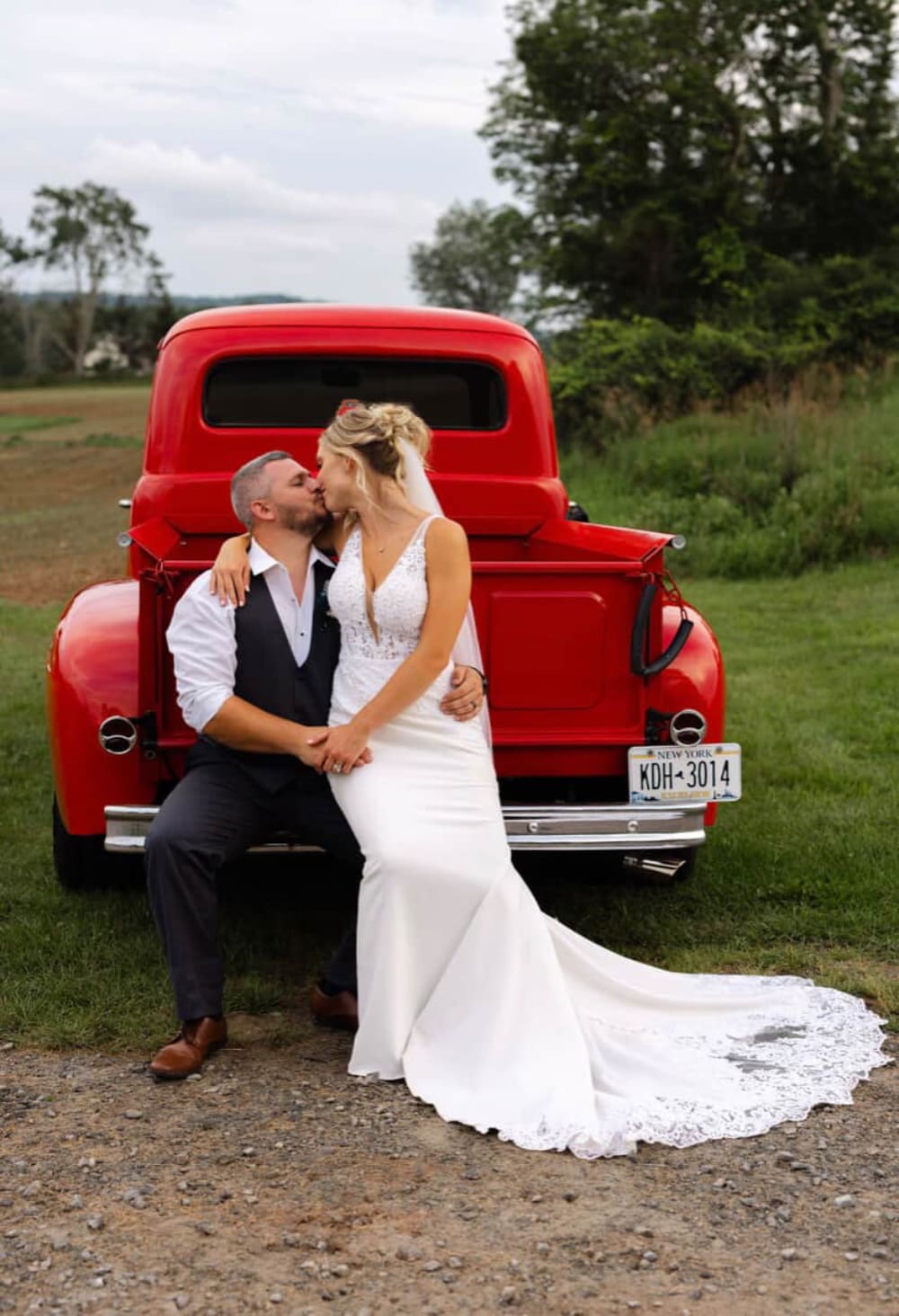 Newlyweds steal a kiss on the tailgate of a vintage red truck at The Barn on Hubbard. Lace train, golden hour light, and all the rustic Catskills charm you could ask for.