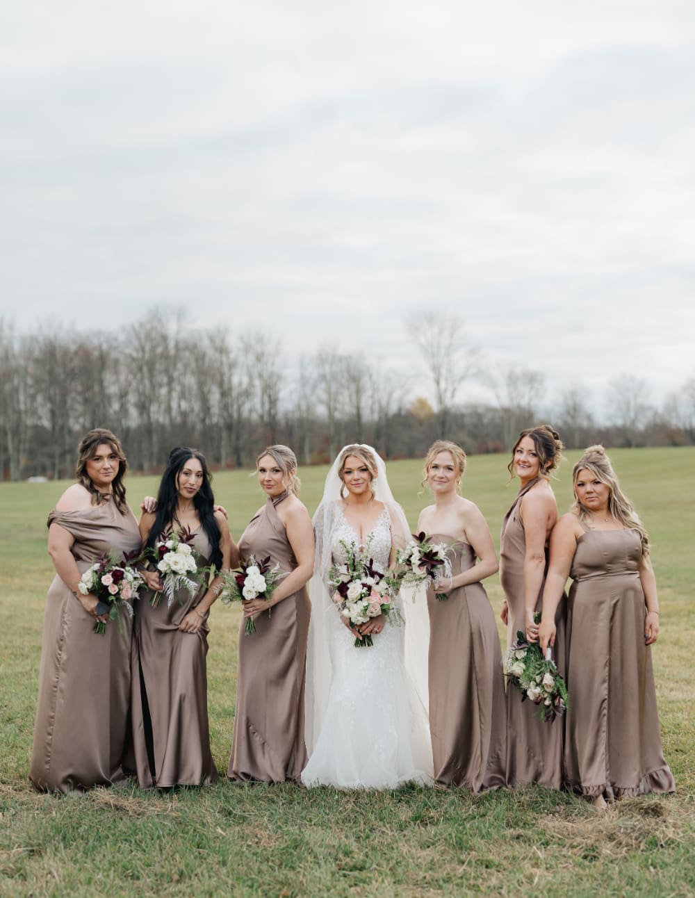 A bride and her bridal party pose on the open fields at The Barn on Hubbard. Coordinated bouquets, matching taupe gowns, and rolling Catskills hills in the background. This is what happens when your wedding vendor list comes together.