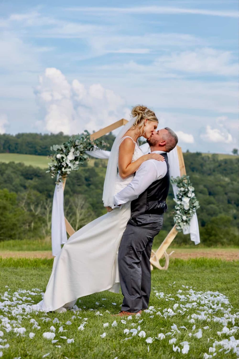 A couple shares their first kiss beneath a hexagonal arch draped in eucalyptus and white florals, with sweeping Catskills views behind them. Ceremony magic at The Barn on Hubbard.