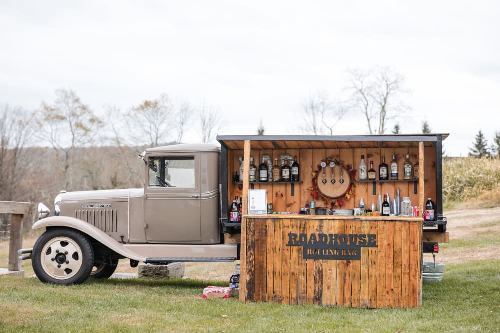 The Roadhouse Rolling Bar set up on the grounds at The Barn on Hubbard. A vintage truck, rustic wood bar, and fall foliage in the distance. This is why a great mobile bar belongs on every wedding vendor list.