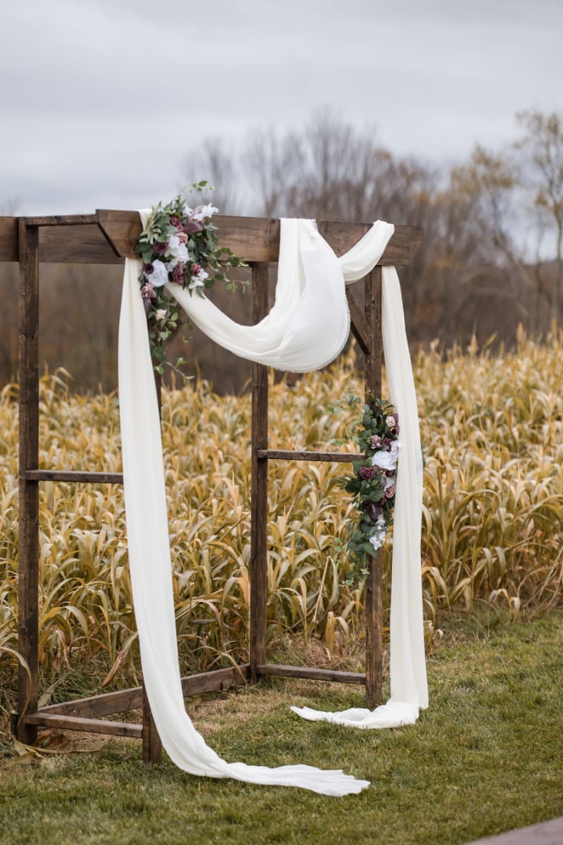 Rustic wedding ceremony arch with flowing fabric and floral accents in an outdoor field setting.