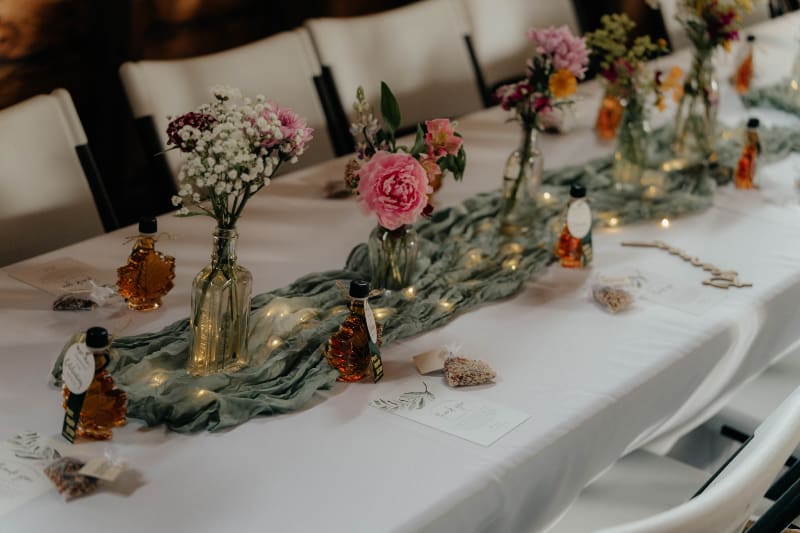 Wedding reception table with pink wildflowers, glass bud vases, sage runner, string lights, and personalized favors.