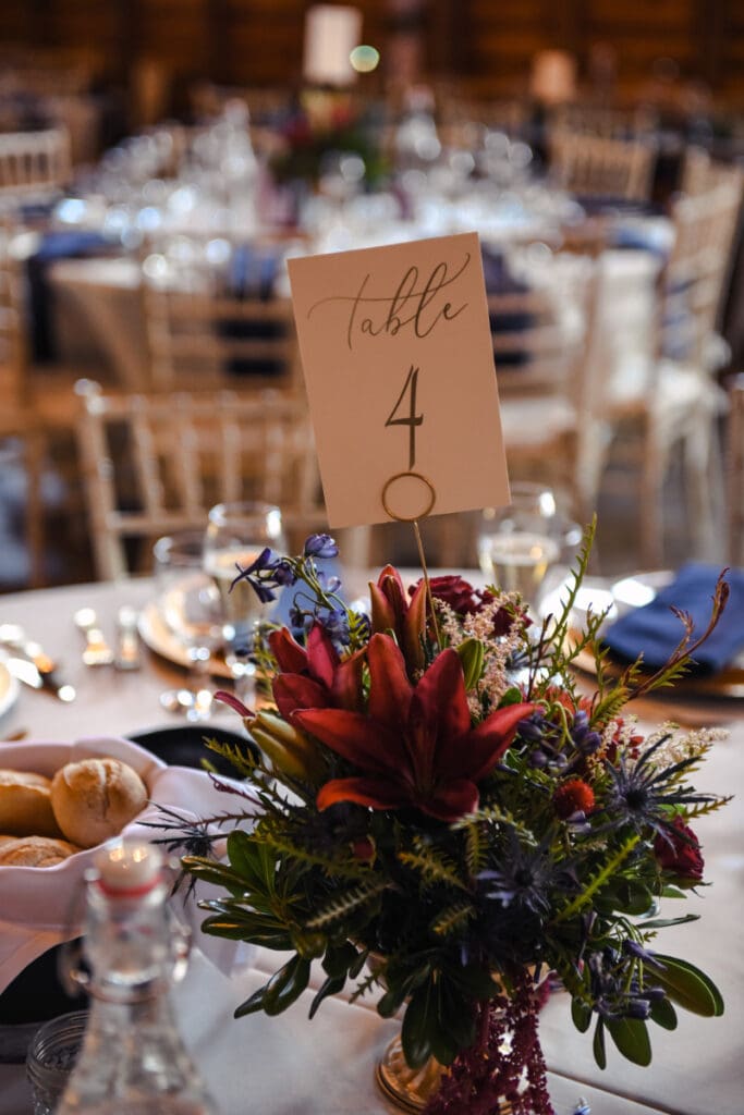 Colorful barn wedding decor ideas with red lilies, greenery, navy napkins, candlelight, and table number four.