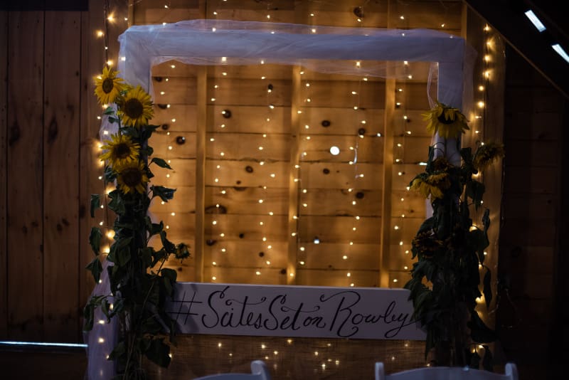 Sunflower-covered photo booth backdrop with warm string lights and a personalized wedding hashtag sign.