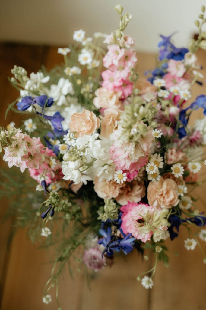 Colorful wedding bouquet with peach roses, pink and white flowers, daisies, blue blooms, and greenery.