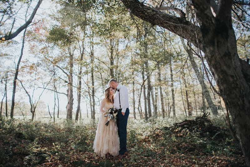 Newlywed couple kisses beneath a large tree in the wooded Catskills landscape at The Barn on Hubbard, highlighting the outdoor photo locations worth considering when preparing questions to ask your wedding venue.
