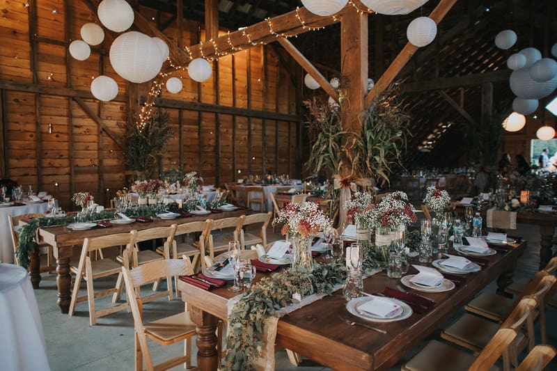 Rustic wedding reception inside The Barn on Hubbard with long wooden tables, flowers, place settings, string lights, and paper lanterns, showing why reception setup belongs on your list of questions to ask your wedding venue.