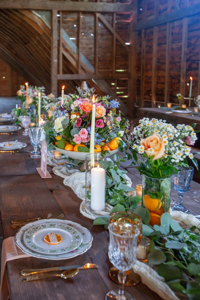 Long wooden reception table inside a rustic barn, decorated with colorful flowers, citrus, greenery, candles, vintage place settings, and glassware.