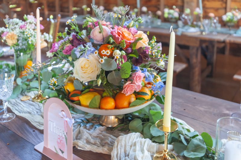 Colorful wedding centerpiece filled with roses, wildflowers, greenery, and fresh citrus, arranged on a rustic reception table with glowing taper candles.