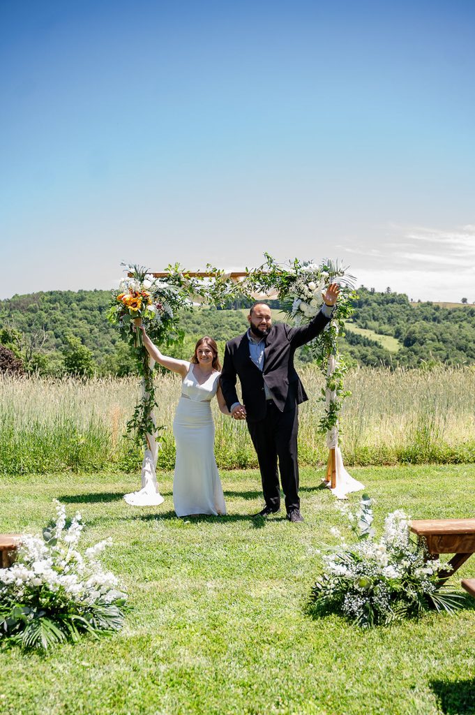 Newlyweds celebrate beneath a flower-covered outdoor ceremony arch, surrounded by rolling hills and countryside views at their fall foliage wedding.