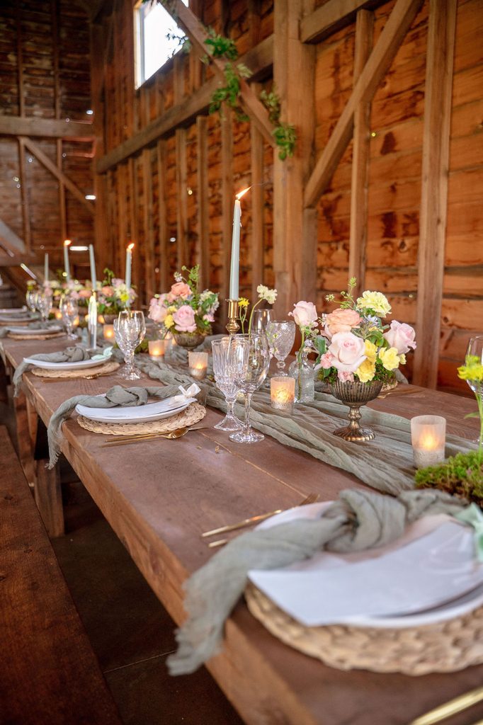 Rustic barn reception table styled with soft pink florals, greenery, taper candles, vintage glassware, woven chargers, and elegant place settings.