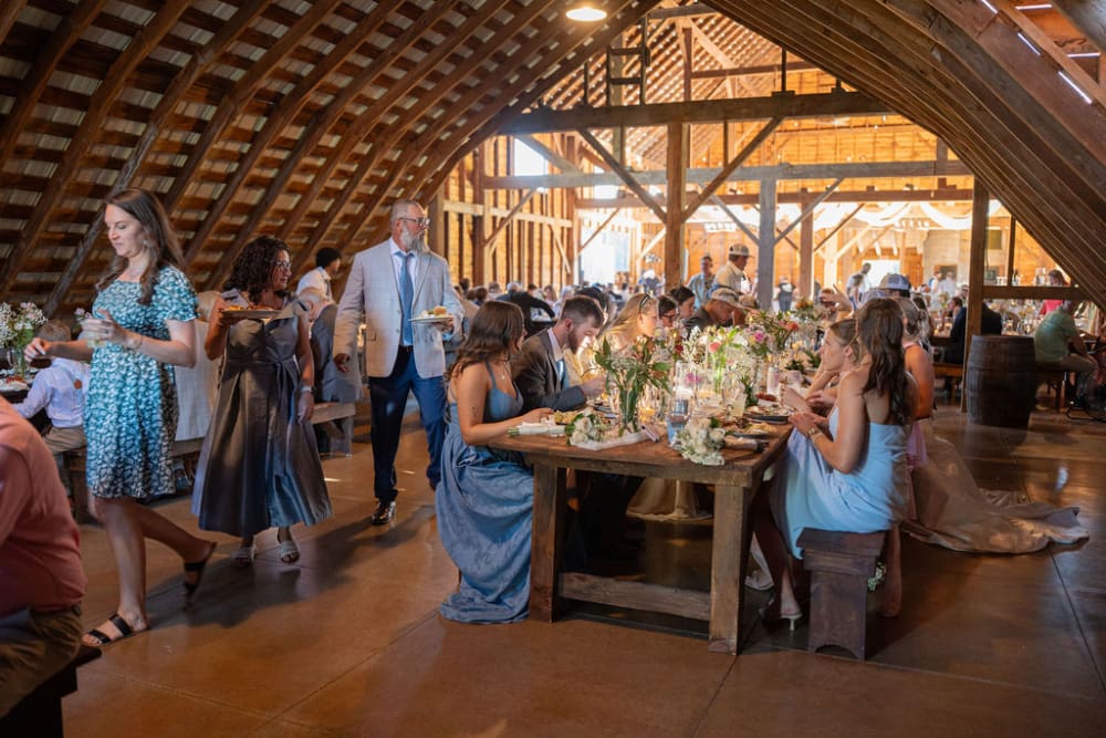 Caterers serve wedding guests at long farm tables with wildflower centerpieces under the arched wooden beams of a restored dairy barn.