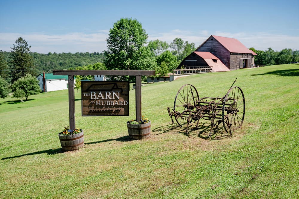 The Barn on Hubbard sign between wine barrel planters, an antique hay rake, and the red-roofed barn on a grassy Callicoon hillside.