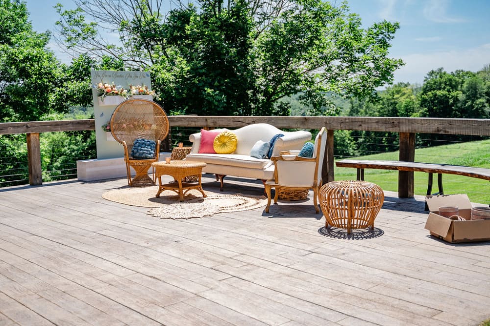 Vintage lounge on an outdoor patio with a white loveseat, bright pillows, a rattan peacock chair, and jute rugs, overlooking trees.
