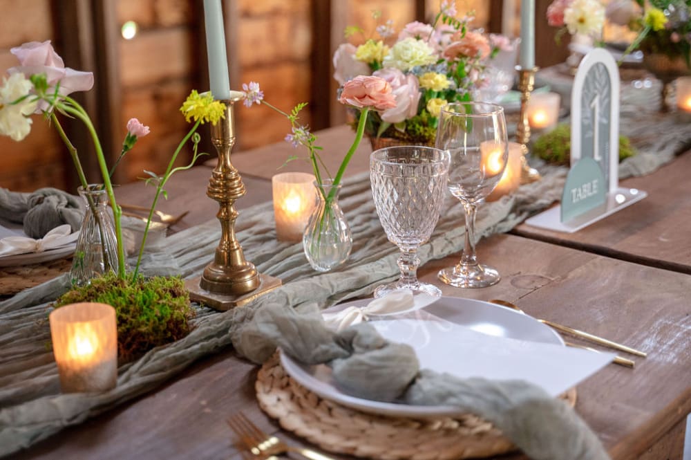 Wedding table with a sage runner, brass candlesticks, glass votive holders, garden roses in bud vases, crystal goblets, and a table number.