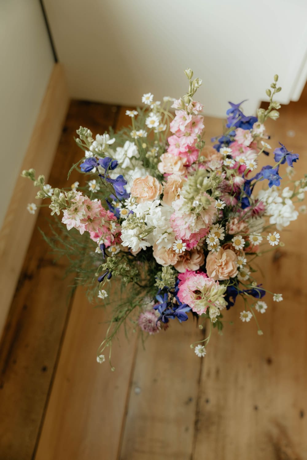 Overhead view of a loose wildflower bouquet with pink and blue delphinium, white stock, peach carnations, and chamomile on wide-plank wood.