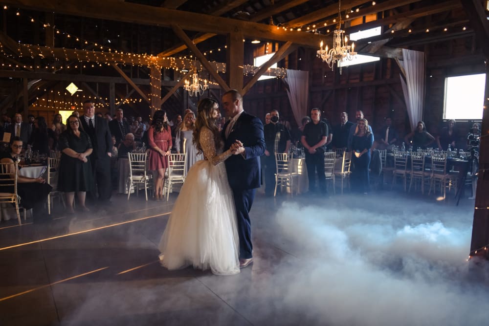 NYC couples can picture this: a bride and groom share their first dance inside The Barn on Hubbard, surrounded by guests, low fog across the floor, crystal chandeliers, string lights, and the barn's original hand-milled wooden beams overhead.