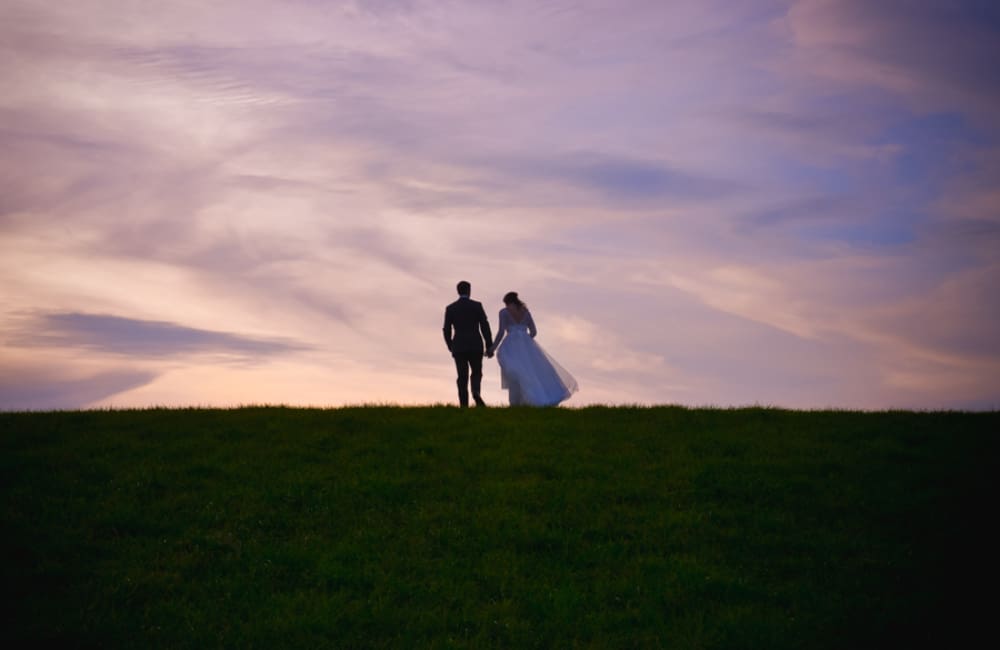A bride and groom hold hands as they walk across the top of a grassy hill, shown in silhouette against a sunset sky of soft pink, lavender, and blue clouds. It's the kind of quiet open space NYC couples come to the Catskills to find.