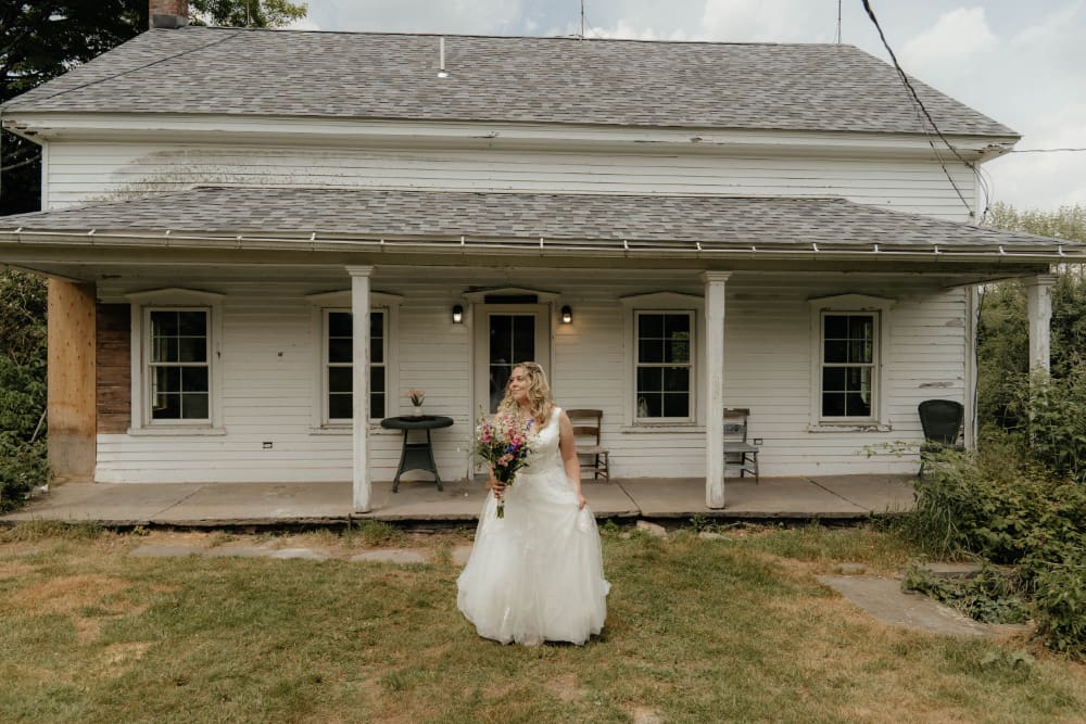 A smiling bride in a sleeveless white lace gown holds a bouquet of pink, purple, and blue wildflowers on the lawn in front of a white farmhouse with a wide covered porch, white columns, wooden chairs, and a small table with fresh flowers.