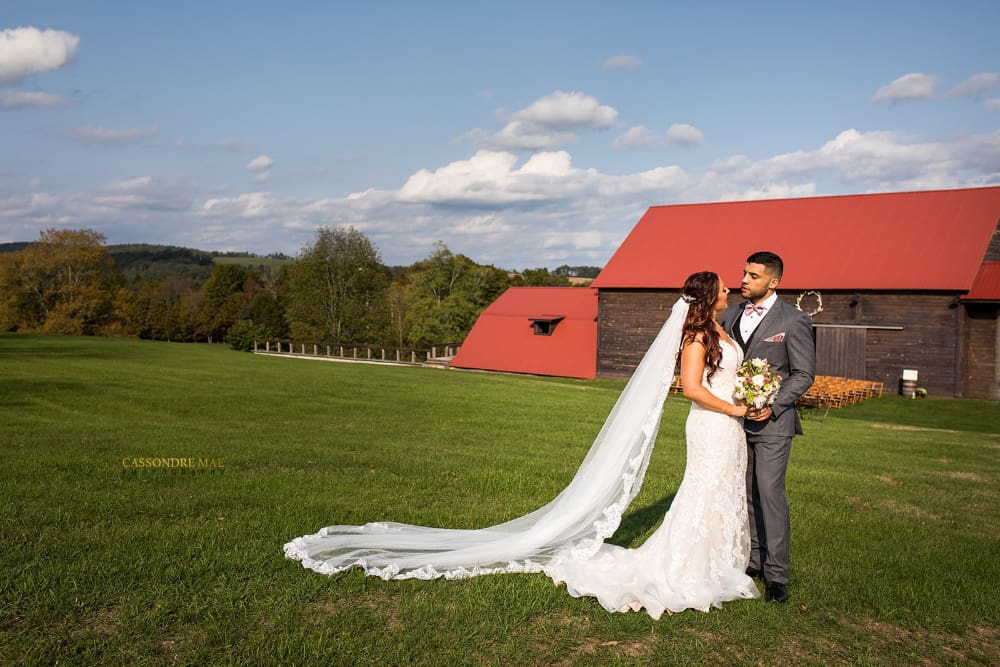 A bride with a long lace-trimmed cathedral veil stands close to her groom on an open lawn in front of the red-roofed dairy barn at The Barn on Hubbard, with ceremony chairs set up beside the barn for NYC couples planning a Callicoon wedding.