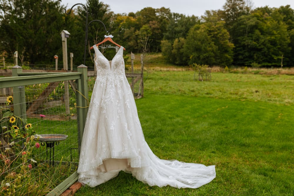 A white beaded A-line wedding gown with a deep V-neckline and long tulle train hangs from a garden hook beside a green wooden fence, with sunflowers, zinnias, and birdhouses nearby and an open green field and trees in the background.