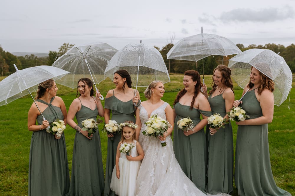 Bride in a lace gown stands with six bridesmaids in sage green chiffon dresses and a young flower girl in white, all holding white rose bouquets and clear bubble umbrellas on a damp green lawn under an overcast sky.