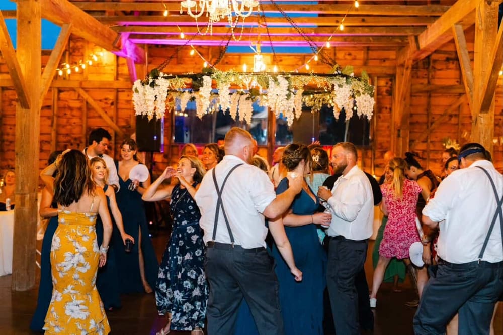Guests dance at a barn wedding reception beneath a hanging hoop of white wisteria and greenery, with string lights, a crystal chandelier, and pink uplighting on the wooden beams. Dancers wear floral dresses and suspenders with white shirts.