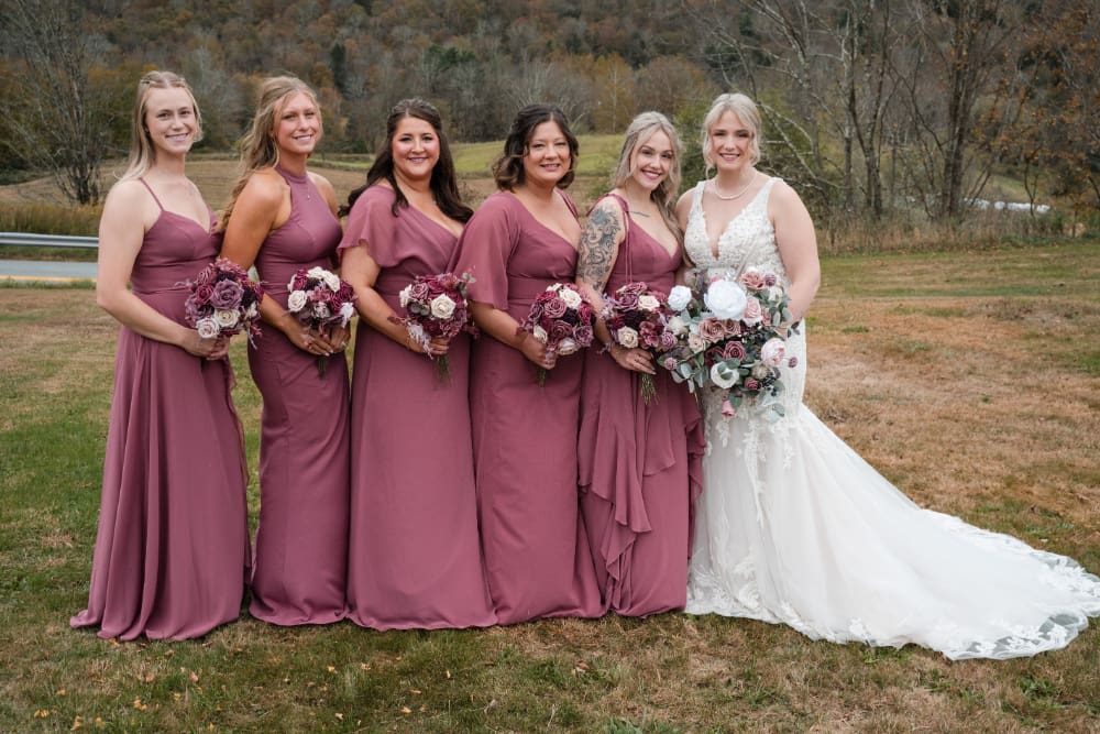 Bride in a lace gown with five bridesmaids in long dusty rose dresses holding mauve and burgundy bouquets, standing on a grassy lawn at a fall wedding with autumn-colored hills and bare trees in the background.