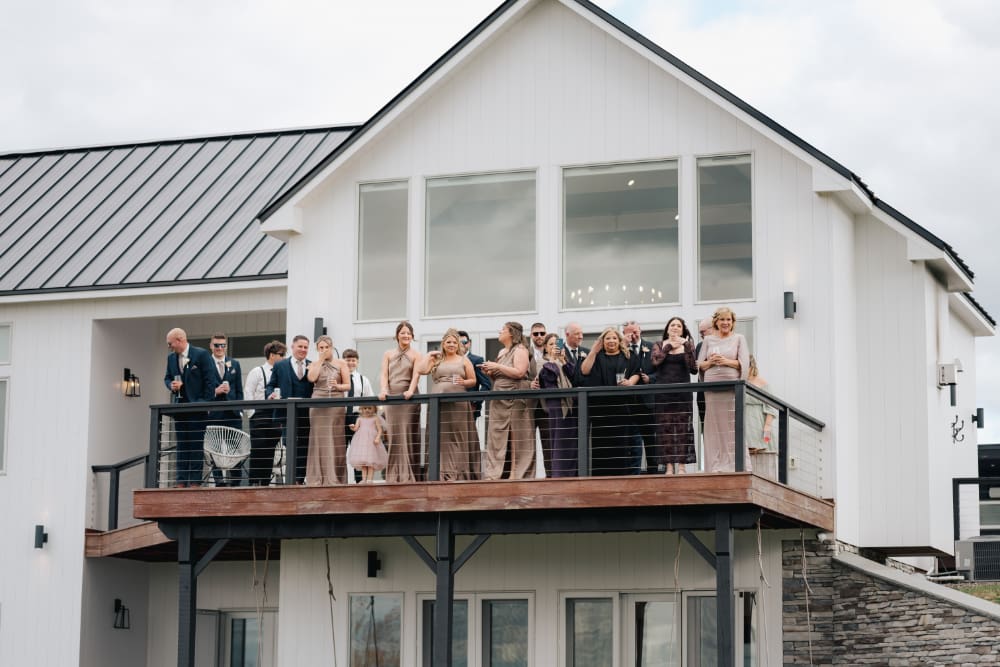 Wedding guests in navy suits and long champagne and plum dresses, plus a little girl in pink, gather along the railing of a modern white farmhouse balcony with tall windows and a metal roof, looking out over the grounds on an overcast day.