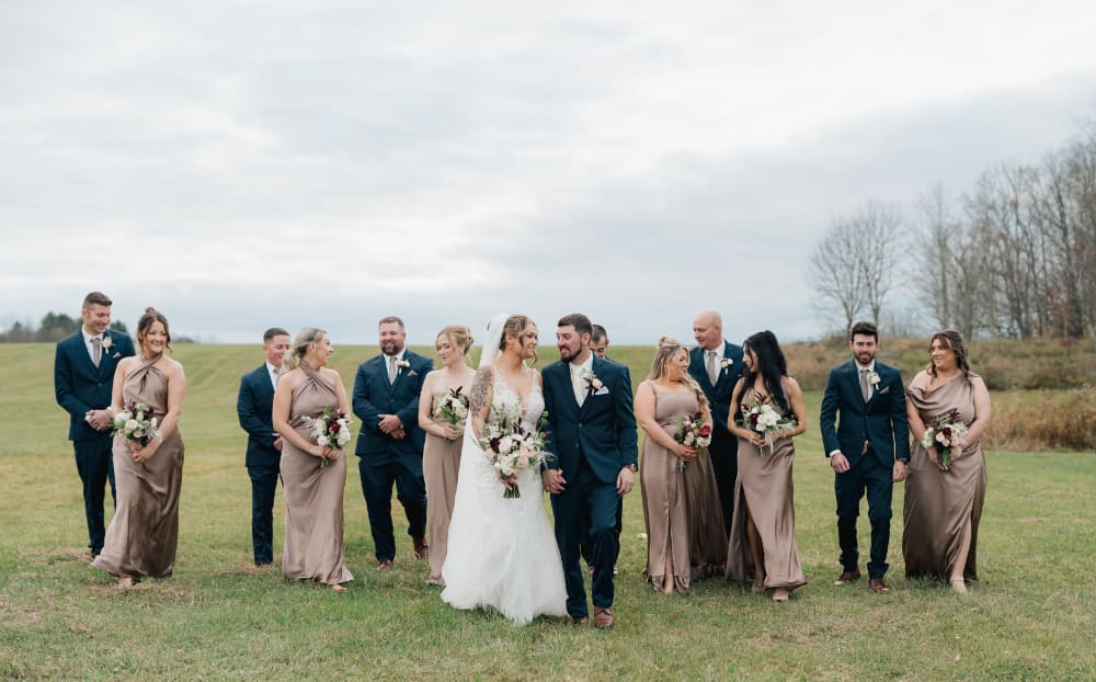 Bride and groom walk hand in hand across a wide grassy field with their wedding party: bridesmaids in satin taupe halter gowns holding burgundy and white bouquets, and groomsmen in navy suits, under an overcast fall sky.