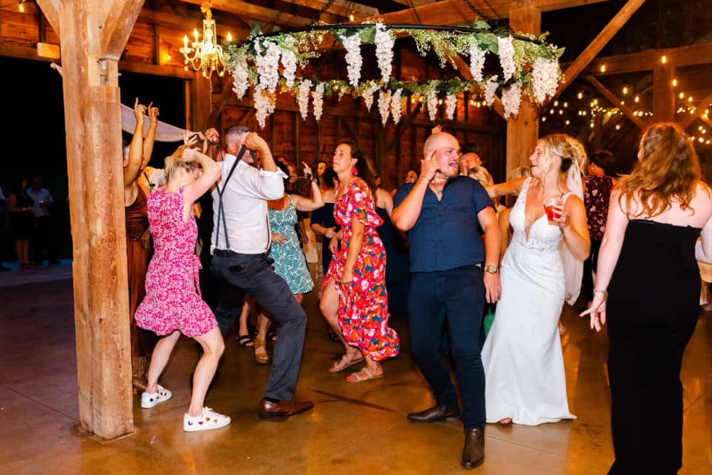 Bride in a fitted white gown dances with guests on the concrete floor of a wedding reception, under a hoop of white wisteria and string lights. Guests wear bright floral dresses, with sneakers and sandals visible on the dance floor.